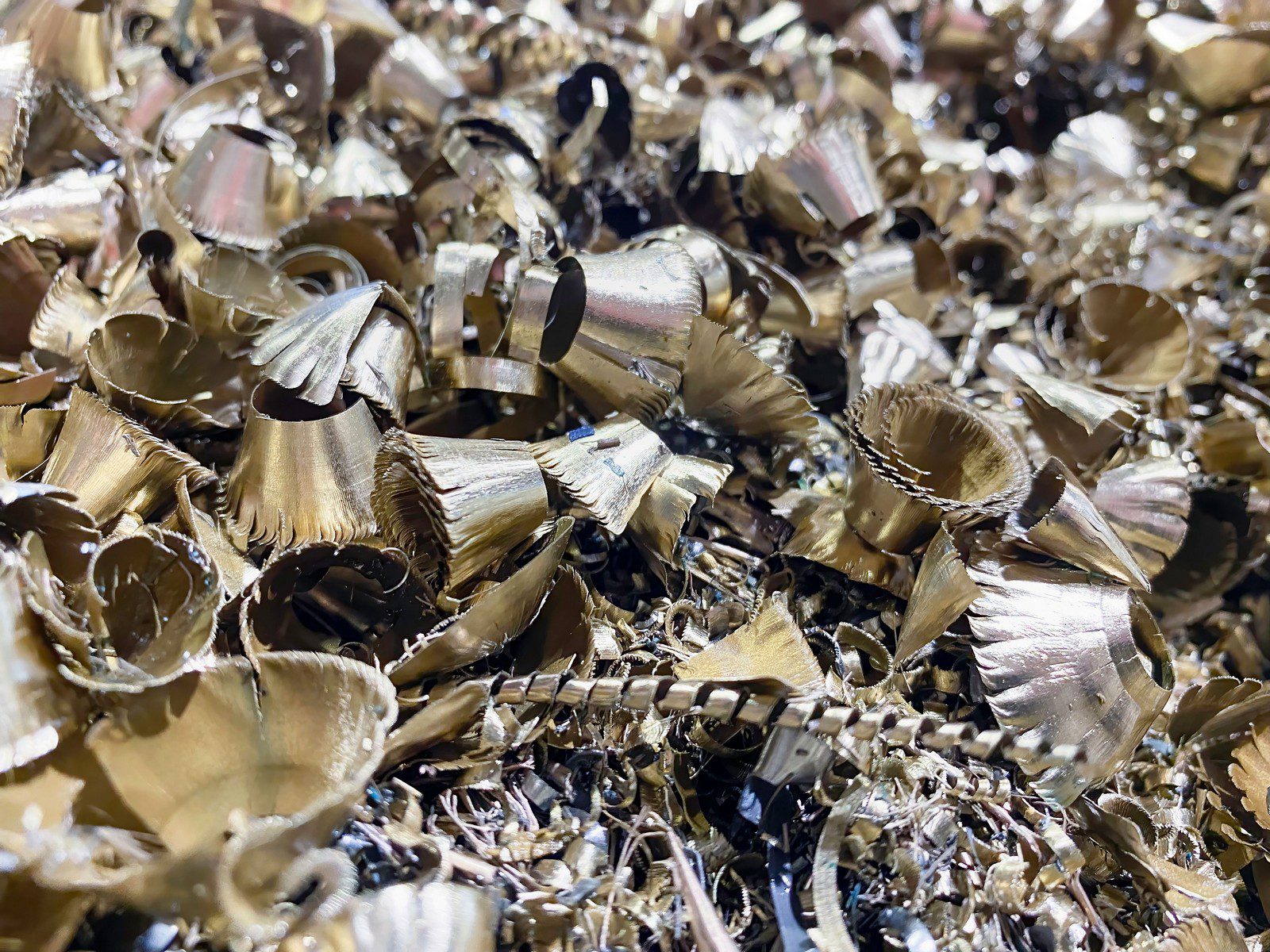 The image shows a pile of metal shavings or swarf. These are the remnants of metal that have been shaved, drilled, turned, or milled from a larger piece during a manufacturing or machining process. The shavings often have a curly or spiral form and can vary in size. They look like they are predominantly made from a material like brass or a similarly coloured metal, given their golden hue. These shavings can be recycled for their metal content.