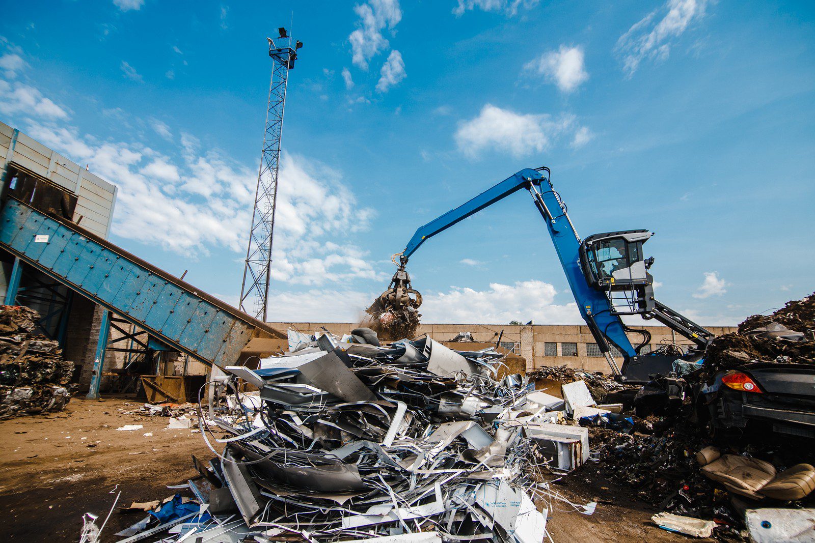 This image shows a material handling machine, likely at a scrapyard or recycling facility. The machine has a grapple attachment and is lifting a load of scrap. The scrap consists of various metallic parts and possibly discarded electronics or appliances, as suggested by the shapes and materials visible. There is a pile of similar scrap materials on the ground. In the background, we can see a tall structure that appears to be a tower, perhaps for lighting or surveillance, and a building with external conveyor structures, suggesting this facility processes the collected materials. The sky is partially cloudy, indicating fair weather conditions.