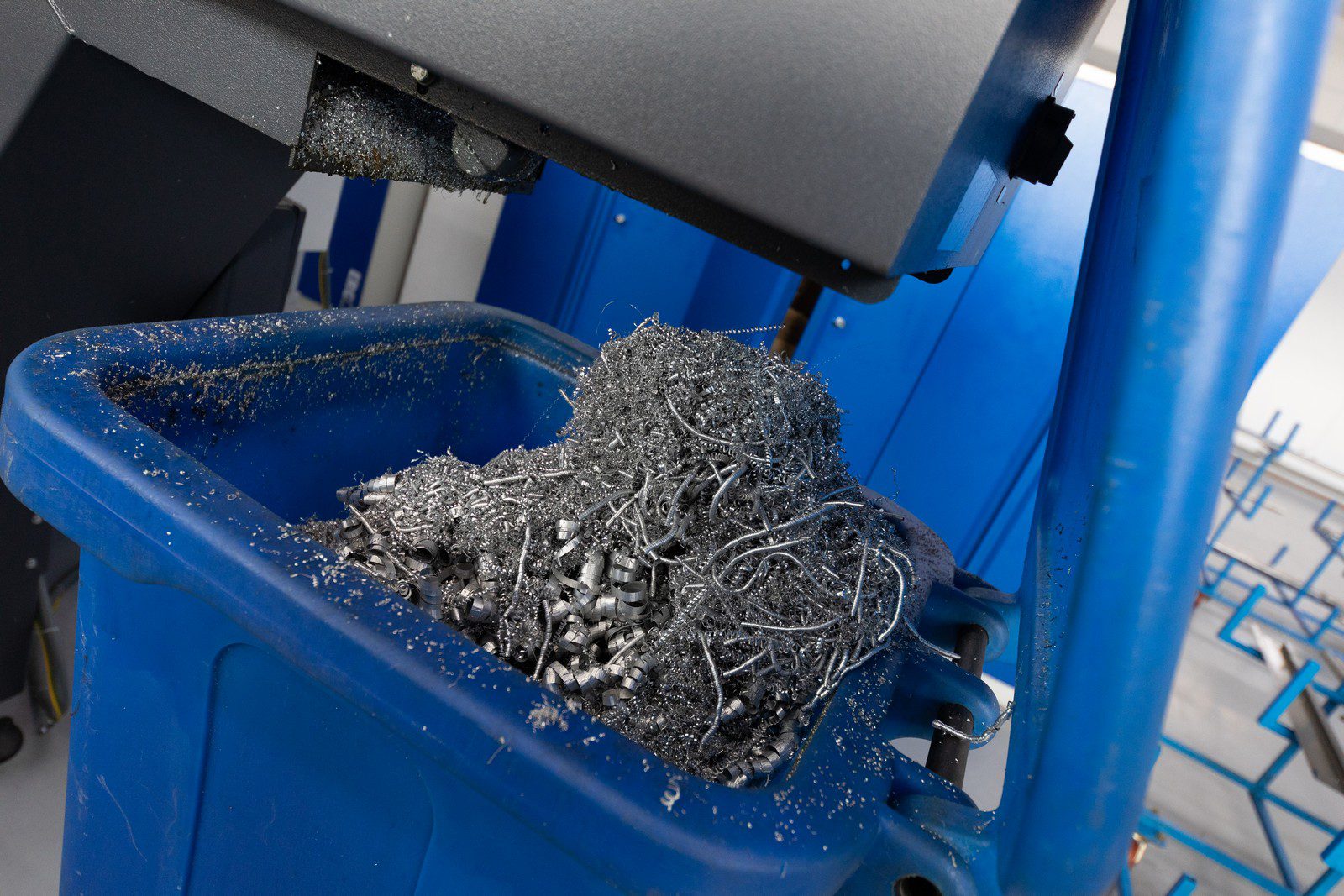 The image shows swarf—metal shavings and filings—accumulated in a blue industrial collection bin. These metal scraps are usually the byproduct of machining operations, like drilling, milling, or turning, where the cutting tools shave off small pieces of metal from workpieces. The environment suggests a workshop or manufacturing environment, where metalworking machinery is used. The pieces of swarf look like they consist of both small, fine particles and larger, curled or spiral shavings, indicative of various cutting processes.