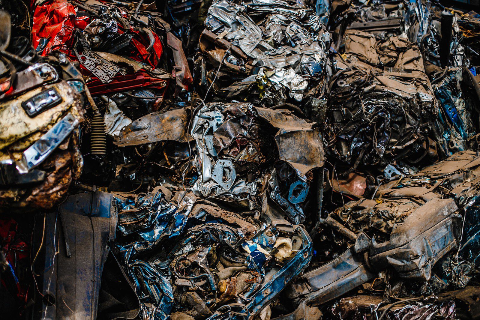 The image shows a pile of crushed and dismantled vehicles, mostly likely in a scrapyard. The different colours and shapes within the pile suggest these are various parts of cars that have been compressed and stacked together. Parts like body panels, frames, and other metallic components can be seen contorted and mangled. This is typically what remains of vehicles after they have been processed through a car crusher, a common step in the recycling process for end-of-life vehicles.