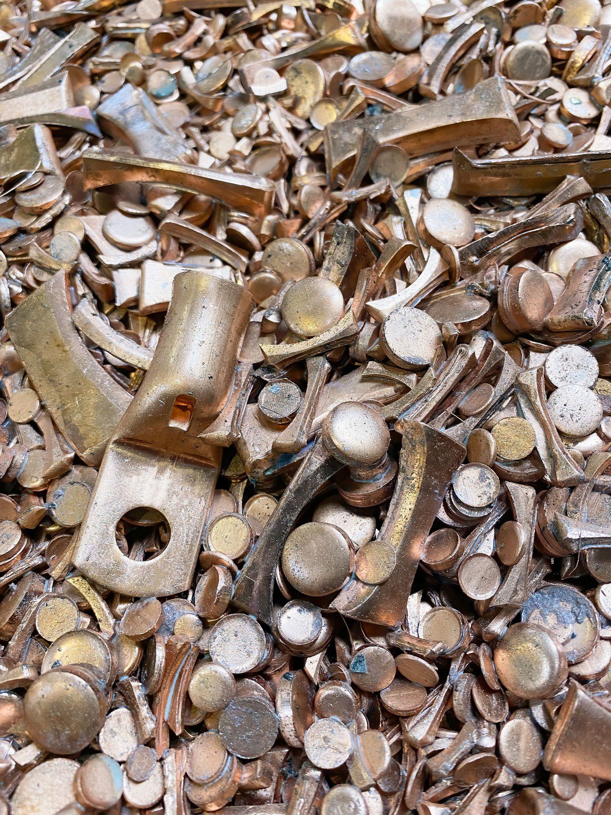 This image shows a heap of various metal objects that appear to be made of brass or a similar alloy. Among the items, there are numerous discarded keys, key shanks, and key bows (the round, flat parts where a keychain would be attached). Additionally, there are unidentifiable small round disks that could potentially be the tops of keys or other components. The pile looks like scrap metal, possibly collected for recycling or remelting. The objects seem worn and used, indicating they might have been gathered after serving their original purposes.