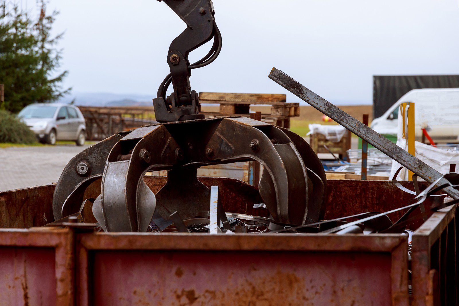 The image shows a large mechanical claw or grapple attached to an excavator arm, positioned over a metal scrap container. The claw is open and positioned as if ready to grab or has just released scrap metal into the container, which is filled with various pieces of metal and debris. In the background, there are outdoor settings that include a white van parked and a wooden structure which might be part of a fence or barrier. Overall, this appears to be a scene from a recycling centre or a construction site where scrap metal is being sorted or processed.