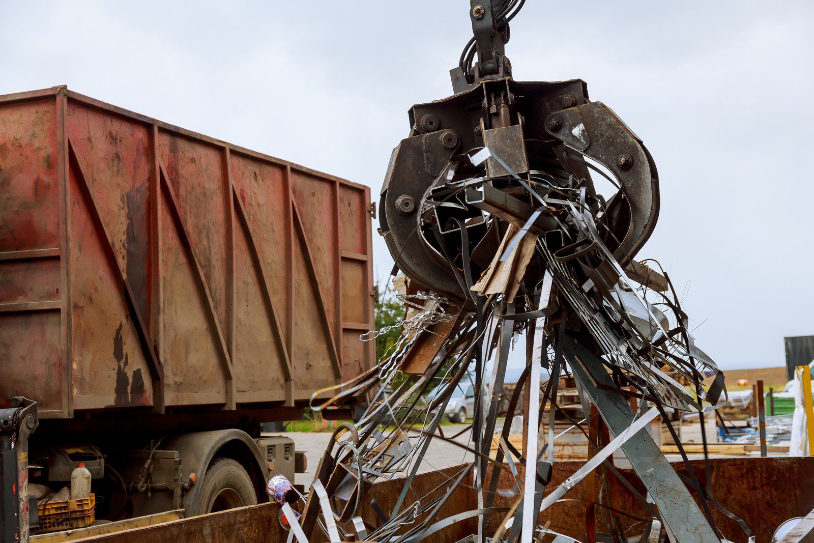 The image shows a scrap metal claw machine grasping various pieces of metal scrap. Above this messy pile of metal parts is the claw, which is part of a larger machine commonly used in recycling facilities to move and sort metal waste. In the background, you can see a large scrap metal container that is likely used to collect the sorted metal before it’s processed for recycling. The machine appears to be in operation at an industrial or recycling site.