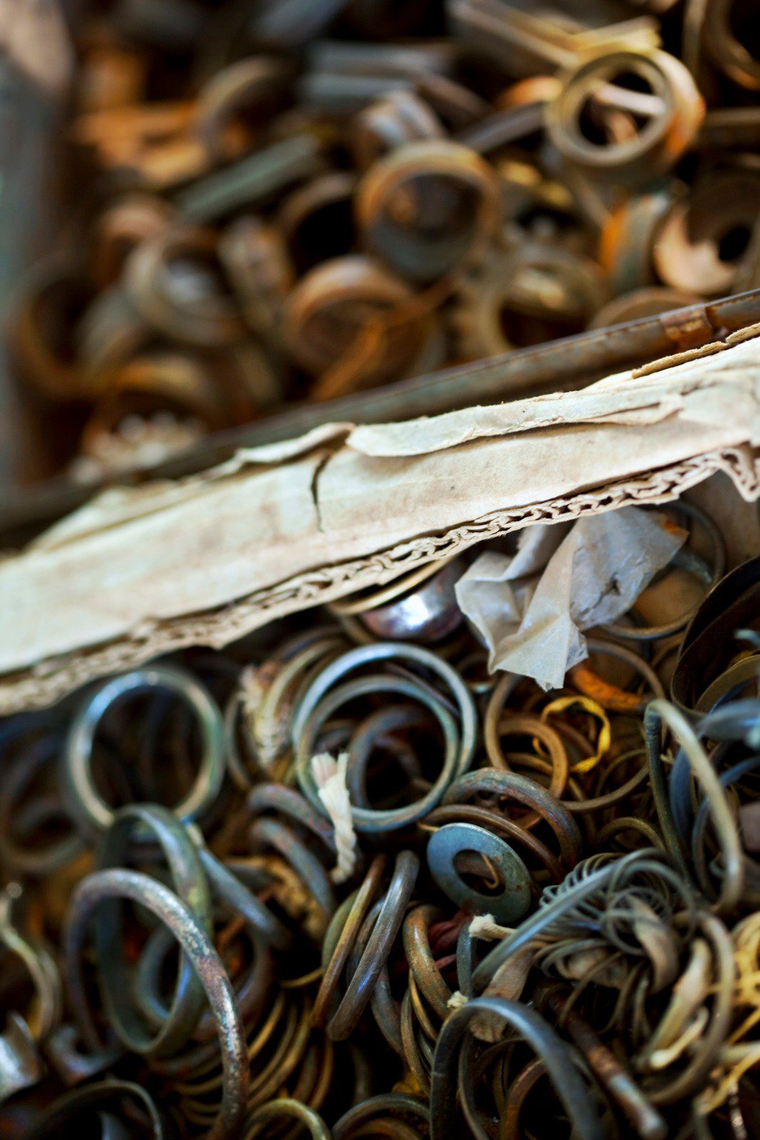 The image displays a collection of various metal rings and objects that appear rusty and aged. They seem to be packed tightly in a container or a compartment. There is also an object that resembles a piece of folded paper or old parchment at the edge, lying over the metal items. The focus is on the foreground, with the background elements being slightly blurred. This creates a depth of field effect that highlights the textures and details of the metal pieces and the paper-like material. The items could be part of a workshop, an antique collection, or materials for artistic or practical use.