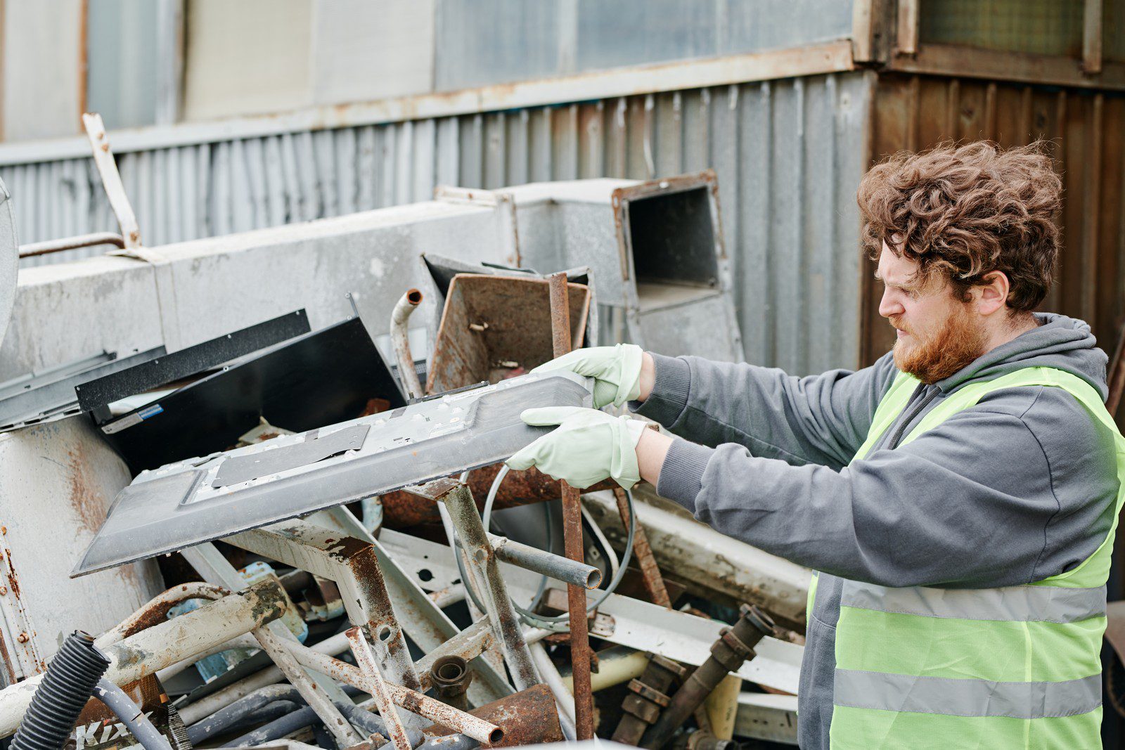 The image shows a person wearing a grey hoodie and a yellow high-visibility vest handling what seems to be a piece of scrap metal. The individual is wearing gloves, suggesting that they are taking precautions while handling potentially sharp or dirty objects. In the background, there are various other metal parts and debris, indicative of a scrapyard or recycling facility where materials are sorted and processed. The person seems focused on the task at hand, which could involve sorting or preparing the metal for recycling.