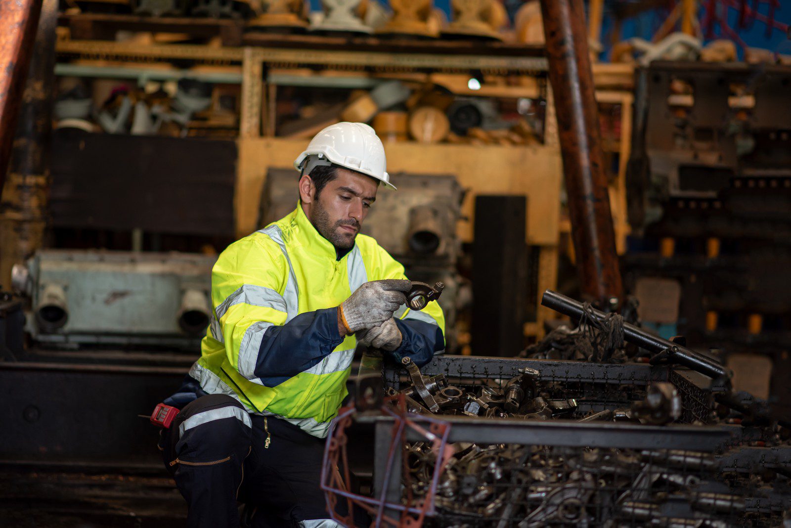 The image depicts a man working in what appears to be an industrial or mechanical workshop environment. He is wearing a high-visibility jacket with reflective stripes, a hard hat, gloves, and work pants, indicating that safety protocols are being followed. The worker is focused on inspecting or working on a component that he is holding in his hands, which could be a part of machinery or equipment.

In the background, there are various pieces of machinery, equipment, and what looks like a disassembled engine or mechanical parts scattered around. The setting suggests a heavy-duty work area, possibly related to manufacturing, construction, or mechanical repairs. The overall impression is that of a professional setting where mechanical or industrial work is being performed.