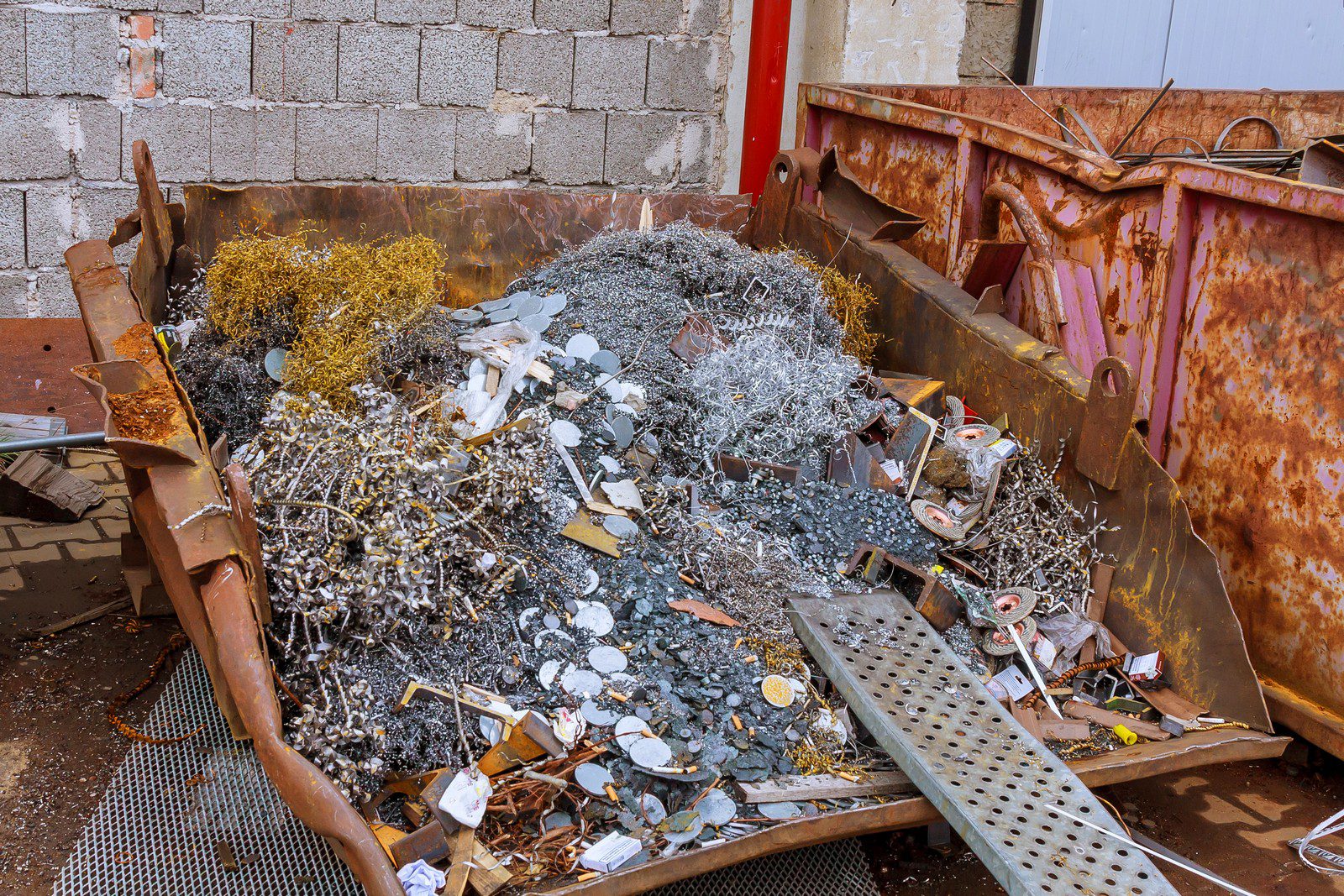 The image shows a large metal dumpster filled with various types of metallic waste and scrap. The contents include items like twisted metal scraps, wires, possibly machine parts, cut pipes, and other miscellaneous metal objects that appear to be collected for recycling or disposal. The rust on the inside of the dumpster suggests that it has been used for this purpose for quite some time. The background shows a wall of cinder blocks, suggesting that the dumpster is located in an industrial area or a recycling facility.