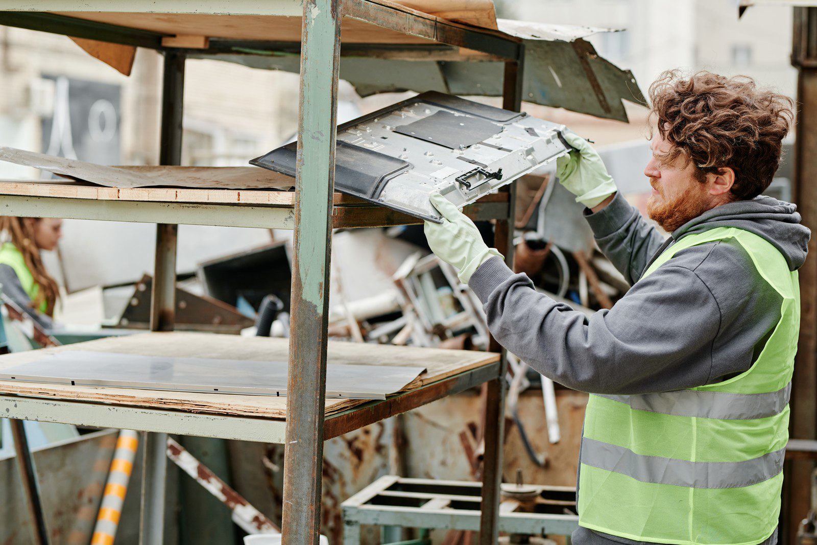 The image shows a man wearing a high-visibility vest and gloves who appears to be working at a recycling or waste disposal facility. He is handling what looks like an electronic component, possibly a piece of a computer or other electronic appliance. There's a wooden structure that might be a sorting station or a container, and various other discarded items can be seen in the background. It seems he's engaged in sorting or processing these items, likely as part of recycling operations.