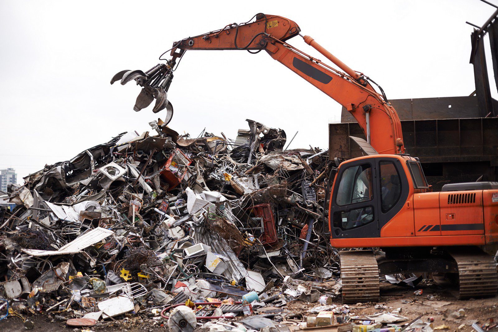 The image shows a large pile of scrap metal and debris being handled by an orange excavator with a grapple attachment. The grapple is open, positioned above the scrap pile as if it's about to grab or release material. The pile contains various types of waste, likely collected for recycling, including crushed vehicles, metal parts, and other unidentifiable components. This is a scene typically found at a scrapyard or recycling facility. There are buildings and a cloudy sky in the background.