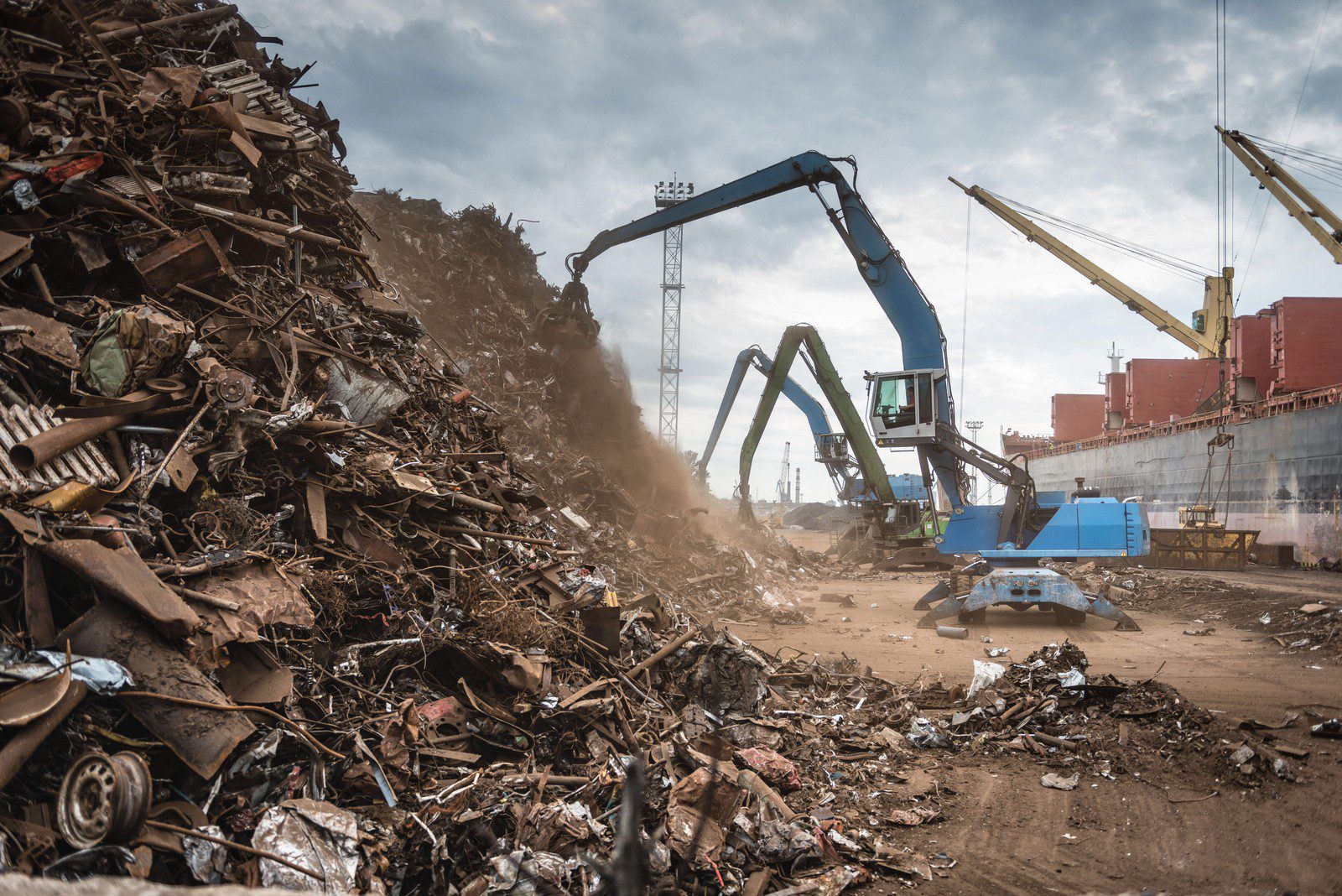 The image shows a scrap metal recycling facility. There is a large pile of various scrap metals including what appear to be parts of vehicles, machinery, and other metal items. In the centre of the image, there's a blue material handler with a grapple attachment, which is commonly used to move and sort scrap material. In the background, there's a large ship docked, suggesting that this facility may be located near a port, and likely the metals could be being prepared for shipment. The sky is relatively clear, indicating the image was captured on a day with good weather conditions.