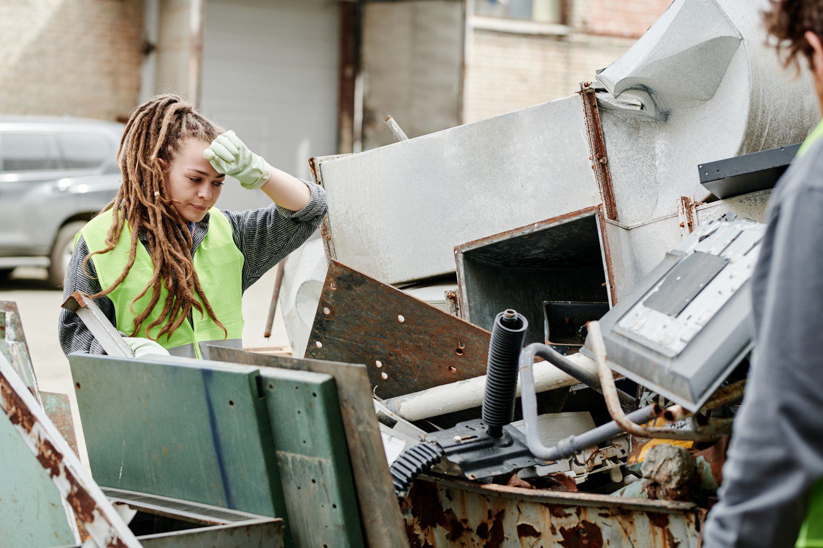 In this image, there is a person with dreadlocks wearing a high-visibility vest and looking into a large industrial waste container. The person appears to be wearing protective gloves and is possibly engaged in waste management or recycling work. The container is filled with various discarded items, including metal parts and other debris. There's a sense that the individual is either sorting through the waste, assessing it for some reason, or maybe about to extract something from it. It looks like an overcast day, and the environment suggests an industrial or semi-industrial area.