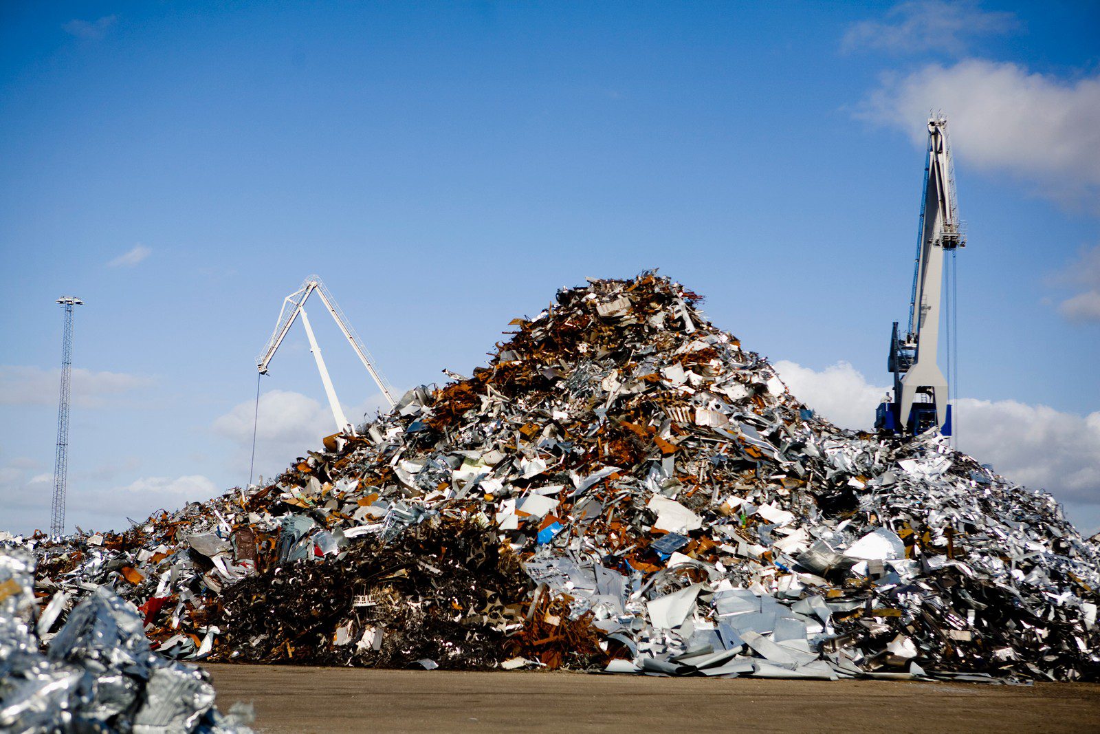 The image shows a large pile of scrap metal and various kinds of waste, possibly at a recycling centre or a scrapyard. There are two cranes in the background, which suggests that the metal is being sorted and moved, likely to be processed and recycled. The sky is clear with a few clouds, indicating that the photo was taken on a day with good weather.