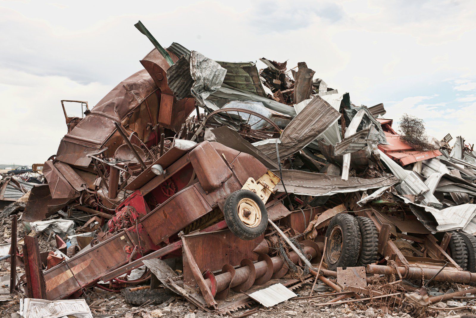 The image depicts a car scrapyard under a cloudy sky, characterized by a cluttered array of partially dismantled and damaged vehicles. The scene is dominated by a multitude of cars in various states of disrepair, stacked haphazardly in piles that tower several vehicles high. The cars, which span a range of makes and models, exhibit signs of rust, missing parts, and broken windows. Some vehicles have hoods or doors ajar, exposing their internal components. The ground is uneven and covered with patches of dirt, debris, and scattered auto parts such as tires, hubcaps, and metallic scraps. In the background, leafless trees are visible against the overcast sky, adding to the desolate and industrial atmosphere of the scrapyard.