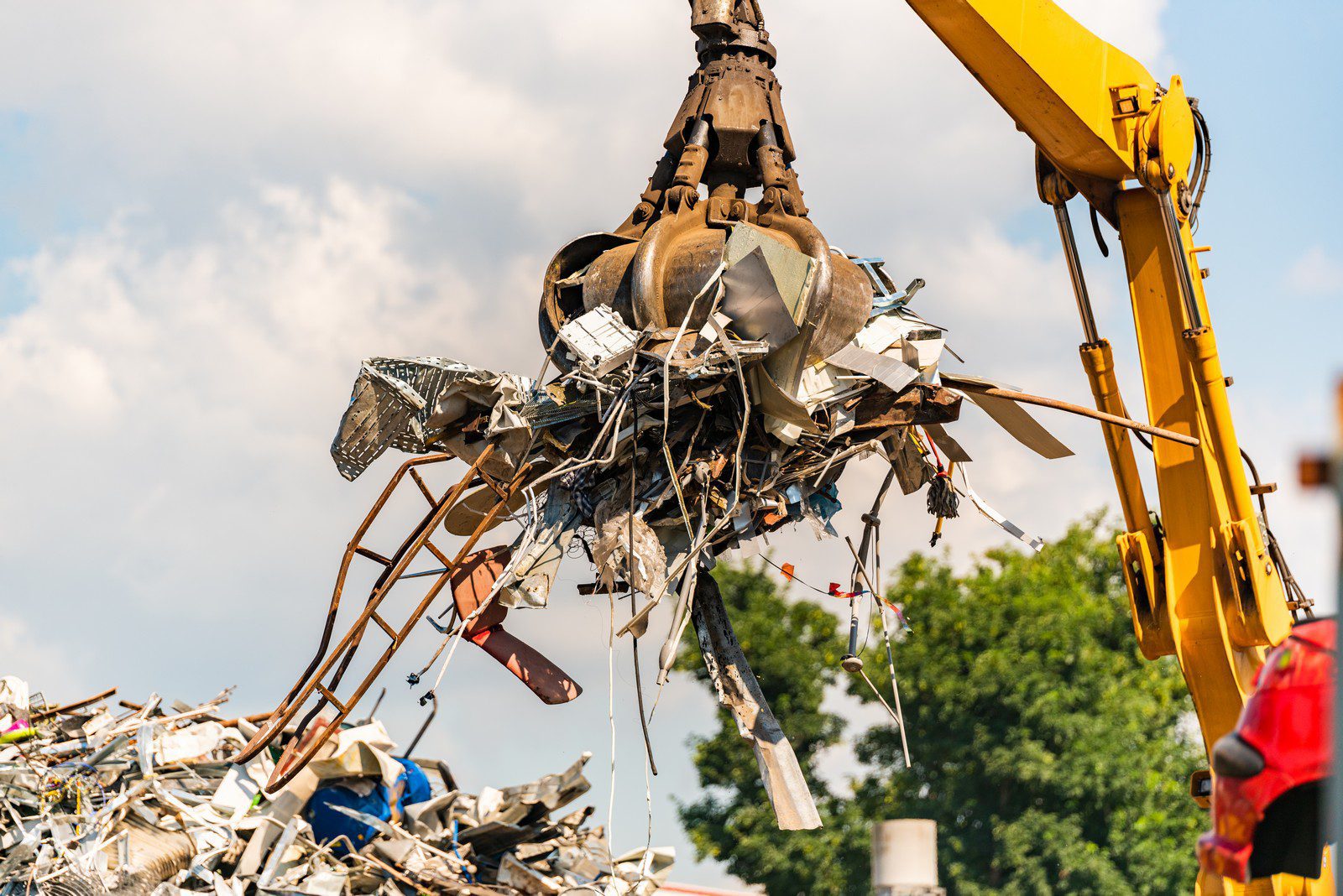 "Close-up of a large industrial crane specialised in recycling metallic waste. The crane's powerful mechanical claw, composed of multiple robust, interlocking metal blades, is prominently featured in the centre of the image. It appears to be in action, gripping a hefty bundle of twisted, rusted metal scraps. The intricate textures of the metallic debris, showcasing a variety of shapes and sizes, create a visually complex scene. In the background, there's a glimpse of an expansive scrapyard piled high with more metallic waste, under an overcast sky, which adds a sense of industrious atmosphere. The dull, weathered hues of the cranes and metal contrast with the subtle sheen of newer pieces, highlighting different stages of wear and oxidation. Overall, the image captures the intensity and scale of industrial metal recycling."