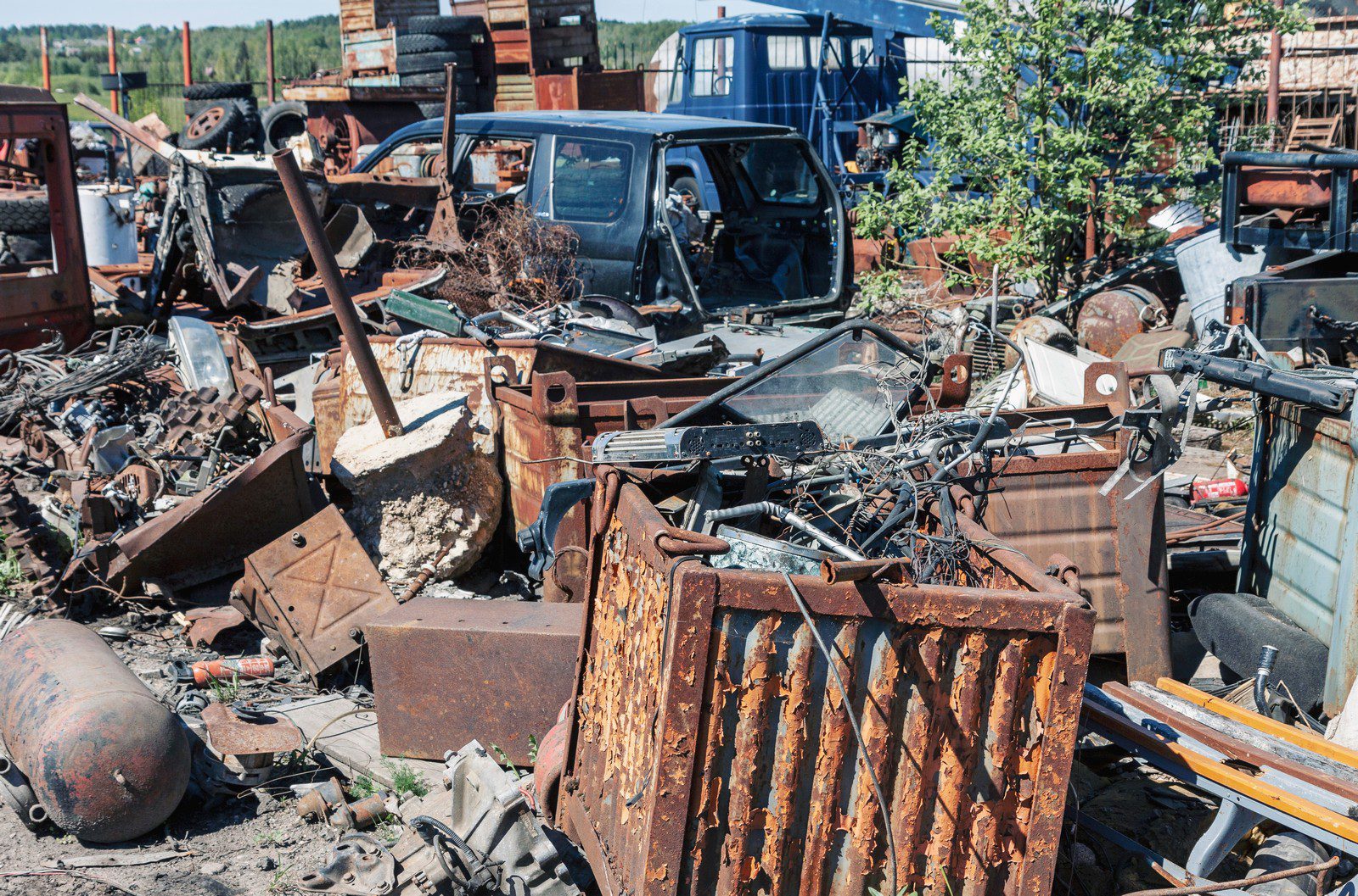 The image depicts a large pile of rusty metal wreckage situated in an outdoor junkyard. The metal debris appears to be an assortment of industrial scrap, including twisted beams, corroded pipes, mangled sheets of metal, and fragments of machinery. The junkyard itself is strewn with similar heaps of metallic rubbish, all characterized by extensive rust and decay. In the background, several dilapidated structures can be seen, which are likely storage units or parts of the junkyard's operational facilities. The sky above is overcast, casting a greyish tone across the scene, which further accentuates the somber and neglected atmosphere of the junkyard. No people or animals are visible in the image, emphasising the desolate nature of the location.