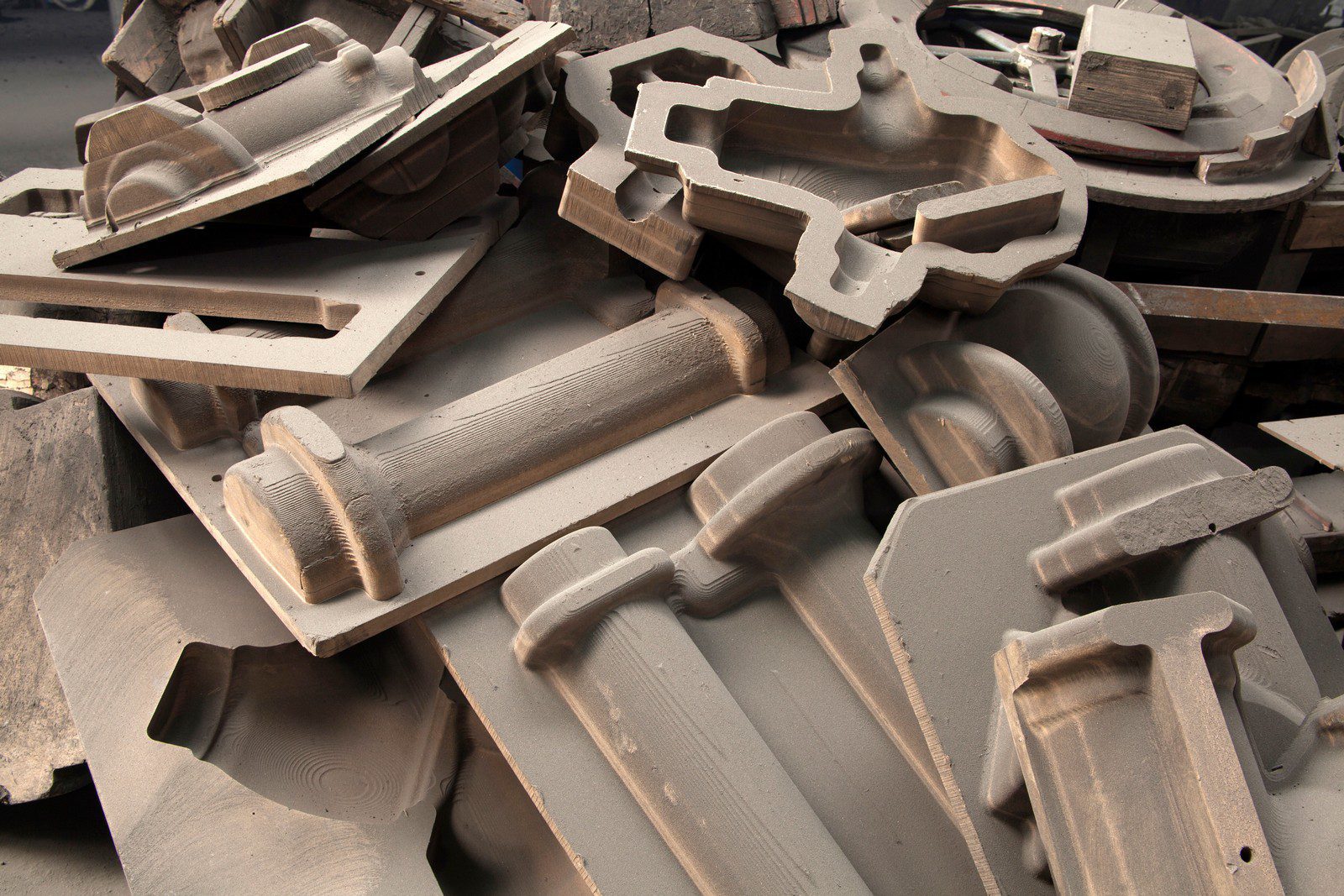 The image displays an industrial indoor setting, specifically a foundry or factory floor dedicated to metal casting. In the foreground, various large and intricately designed metal castings are prominently positioned, showcasing the complex shapes and details produced. The metal pieces exhibit a dull, metallic sheen, reflecting the harsh, artificial lighting typical of industrial environments. Surrounding the castings are scattered tools, molds, and machinery used in the casting process, indicating an active workspace. The background features a series of heavy-duty industrial equipment, including furnaces, conveyor belts, and large containers, all integral to metal casting operations. The factory setting is somewhat cluttered, with various materials and work-in-progress items placed haphazardly, suggesting a busy and productive atmosphere. The walls of the factory are lined with electrical conduits and safety signage, emphasising the industrial nature and focus on operational safety. The overall ambiance of the image is that of a robust industrial environment where precision and craftsmanship in metalwork are on display.